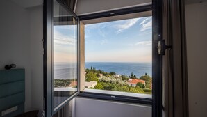 A room with a large window overlooking a coastal landscape, a bed, and a teal-colored headboard.