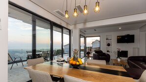 A modern dining area with a wooden table, grey chairs, and a fruit bowl. There is a large window with a view of the ocean and a flat-screen TV mounted on the wall.