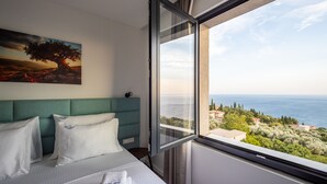 A hotel room with a bed, a headboard, a painting, and a window with a view of the sea and buildings.