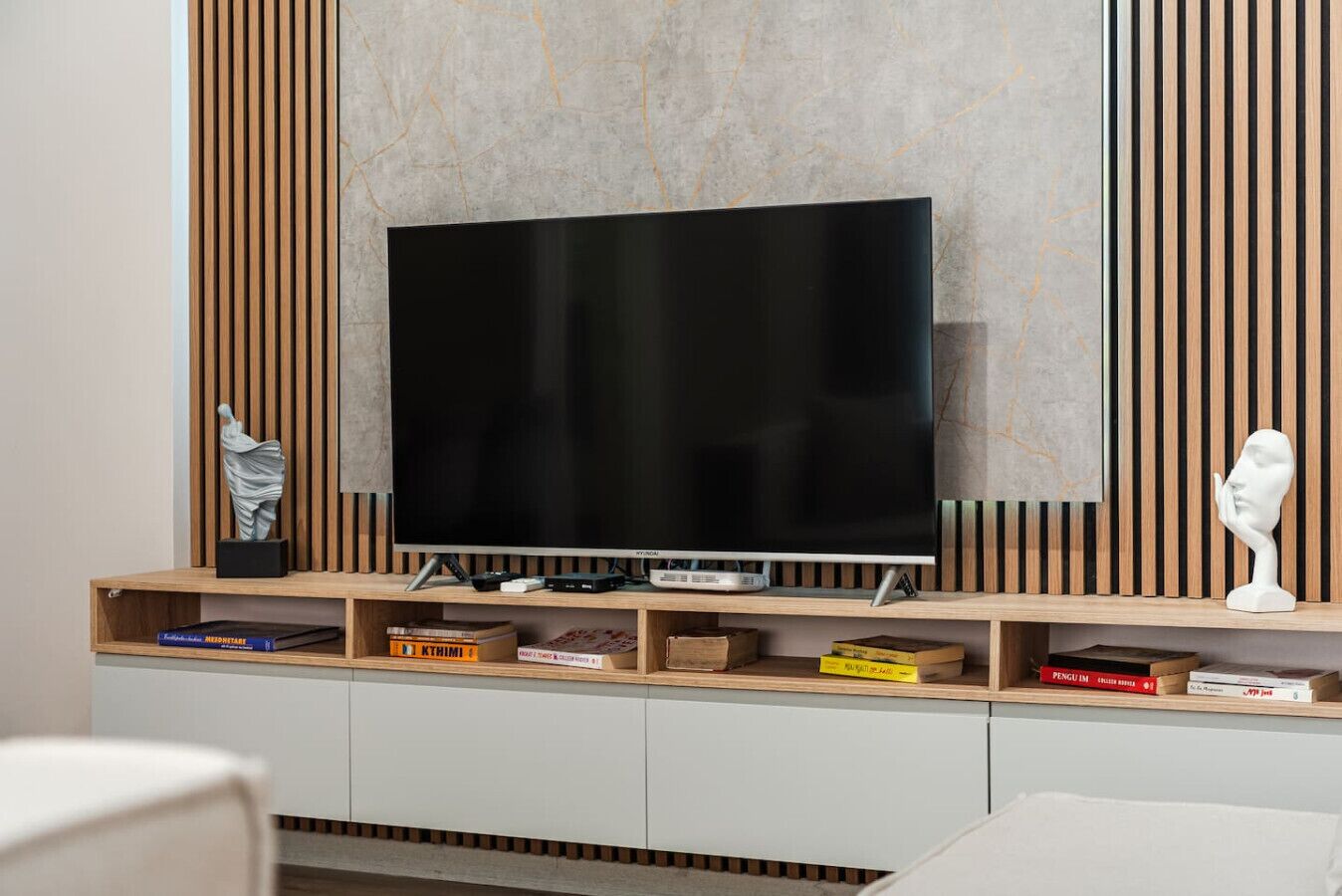 Ein modernes Wohnzimmer mit Flachbildfernseher auf einem Holz-TV-Möbel, Büchern und zwei Skulpturen.