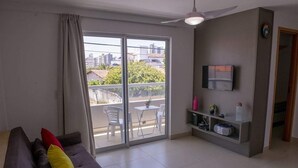 Ein Wohnzimmer mit einer Couch, einem an der Wand montierten Fernseher, einem Balkon mit Tisch und Stühlen sowie einem Deckenventilator.