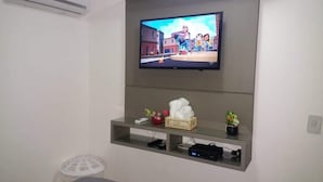 Ein Zimmer mit an der Wand befestigtem Fernseher, einem Regal mit Korb, einer Fernbedienung und einer kleinen Pflanze.