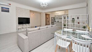 Una sala de estar moderna con un sofá grande de color gris, un televisor de pantalla plana y una zona de comedor con una mesa de vidrio y sillas blancas.