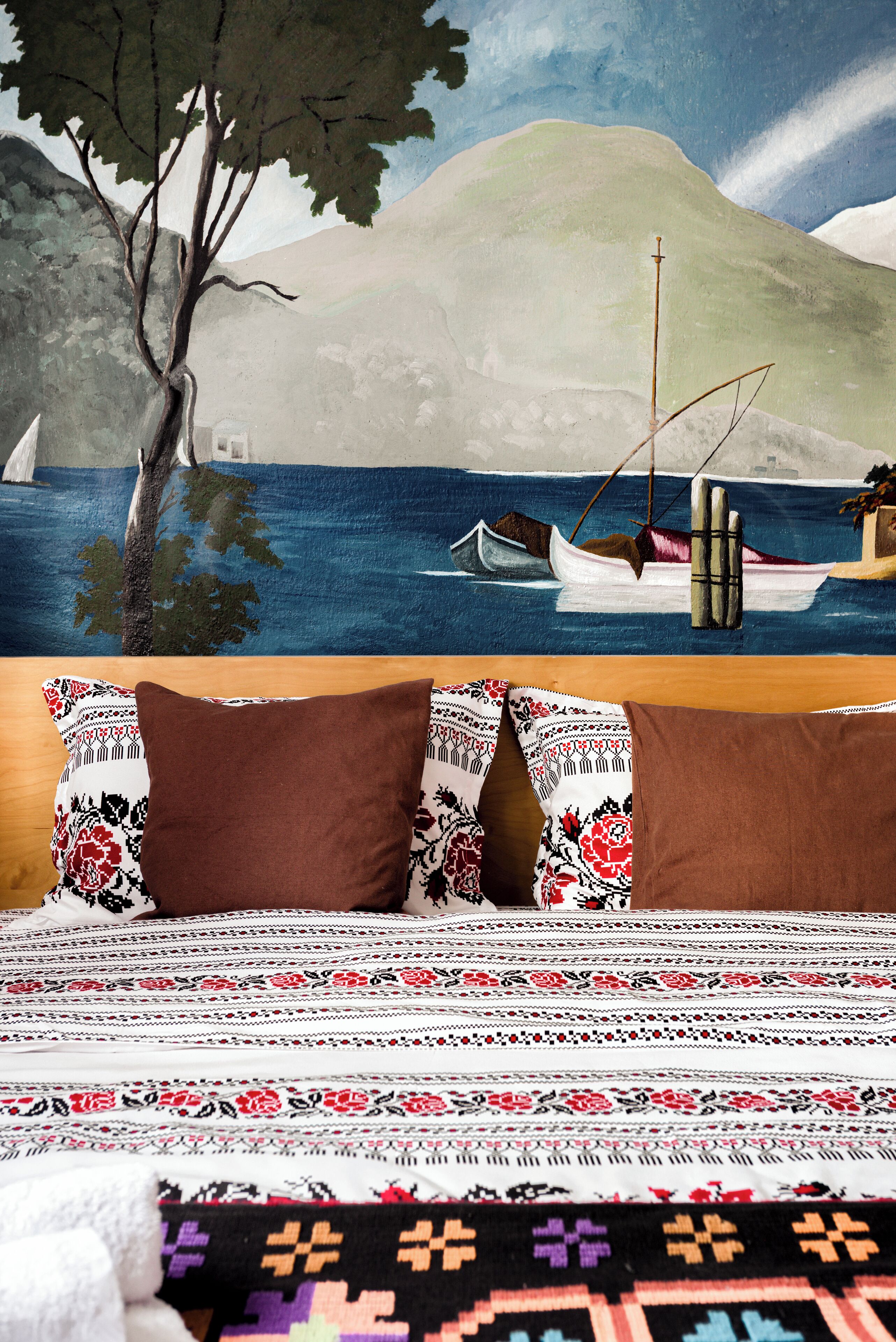 A bed with a patterned bedspread and two pillows, a wall with a painting of a coastal scene, and a wooden headboard.