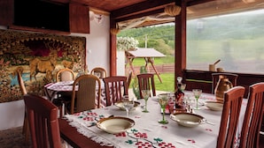 A dining area with a table set for four, surrounded by wooden chairs, a tapestry, and a view of a green landscape. - Transylvanian Home - Mountain View, BBQ & Pool (Bratca)