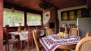 A rustic dining area with wooden tables and chairs, a brick fireplace, and a large window overlooking greenery. - Transylvanian Home - Mountain View, BBQ & Pool (Bratca)