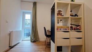 Une pièce avec un plancher en bois, un radiateur, une fenêtre donnant sur une clôture, un bureau avec une chaise et une bibliothèque remplie de jouets et de livres.