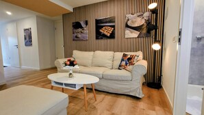 Un salon moderne avec un canapé beige, une table basse ronde et une petite table d’appoint sur laquelle est posé un vase de fleurs. Le sol est recouvert de parquet et un mur est décoré d’un papier peint à rayures.