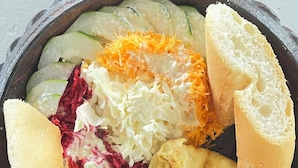 A bowl of food with various items including bread, cucumbers, shredded cheese, and coleslaw.