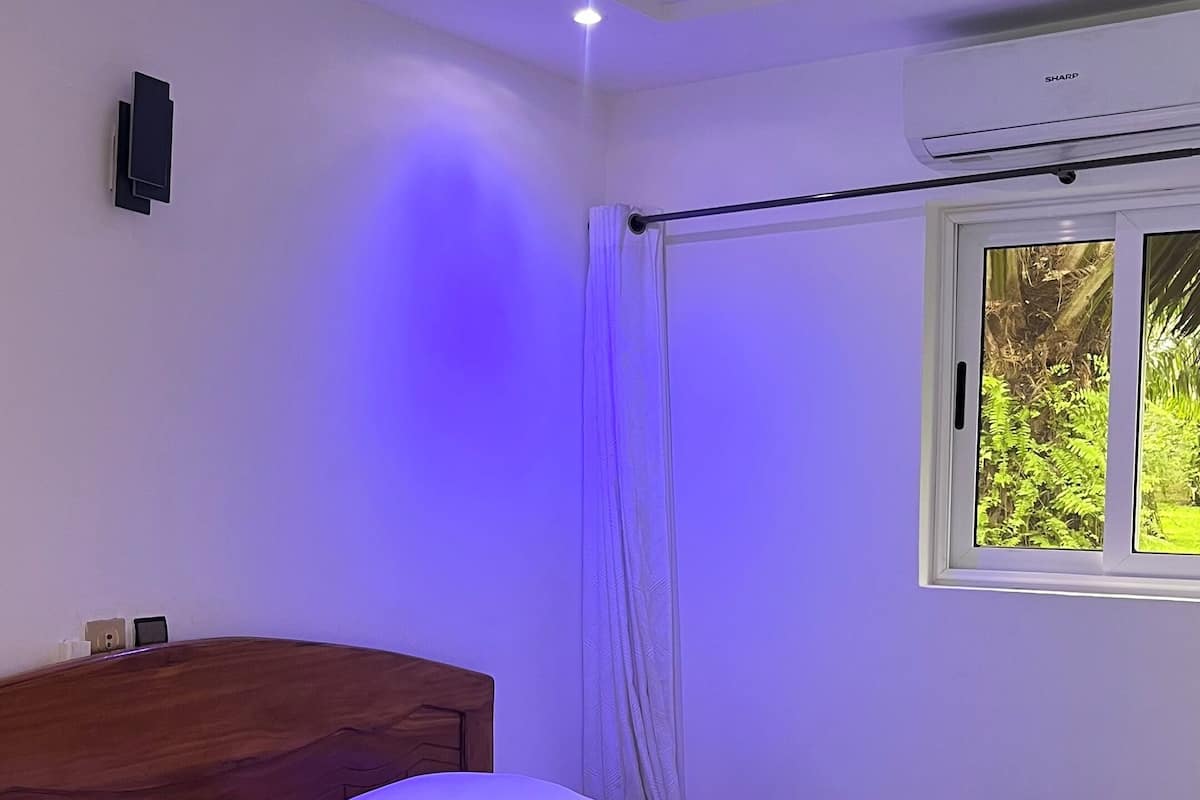 Una habitación con cama, cabecera de madera, ventana con vista a follaje y equipo de aire acondicionado.