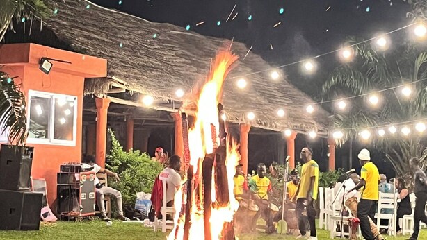 An outdoor event with a central fire, people seated on chairs, and a covered area with string lights.