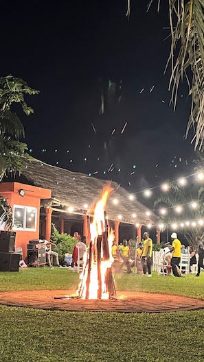 Eine Outdoor-Veranstaltung mit einem zentralen Lagerfeuer, Personen, die auf Stühlen sitzen, und einem überdachten Bereich mit Lichterketten.