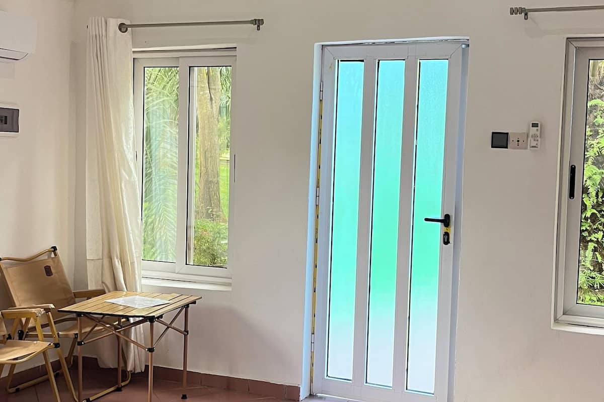 Un dormitorio con una cama, una mesa de madera y una silla. Hay una ventana con cortinas, una puerta con manija y un estante con un dispositivo.