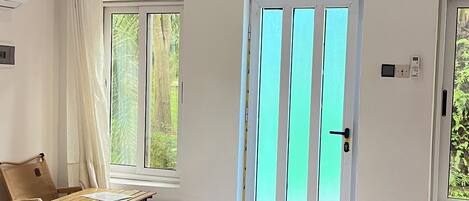 Un dormitorio con una cama, una mesa de madera y una silla. Hay una ventana con cortinas, una puerta con manija y un estante con un dispositivo.