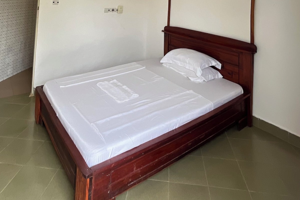 Un dormitorio con una cama de madera, ropa de cama blanca, ventilador de techo, luces empotradas y suelo de baldosas.