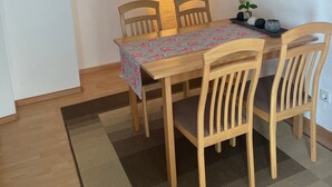 Un comedor con una mesa de madera, cuatro sillas, un mantel floral, una bandeja con objetos decorativos, un cuadro colgado en la pared y una lámpara de pie.