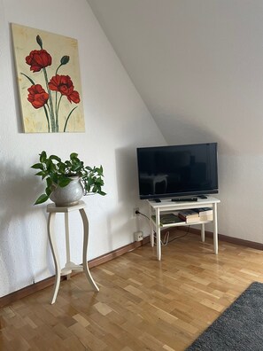 Une pièce avec un téléviseur posé sur un meuble blanc, une plante en pot sur une petite table et un tableau représentant des fleurs rouges accroché au mur.