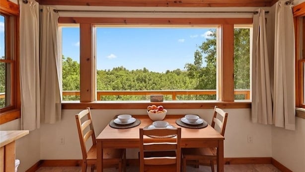 A dining area with a wooden table and chairs, a bowl of fruit, and a view of a forest through large windows. - Your Quiet Mountain Escape (Boone)