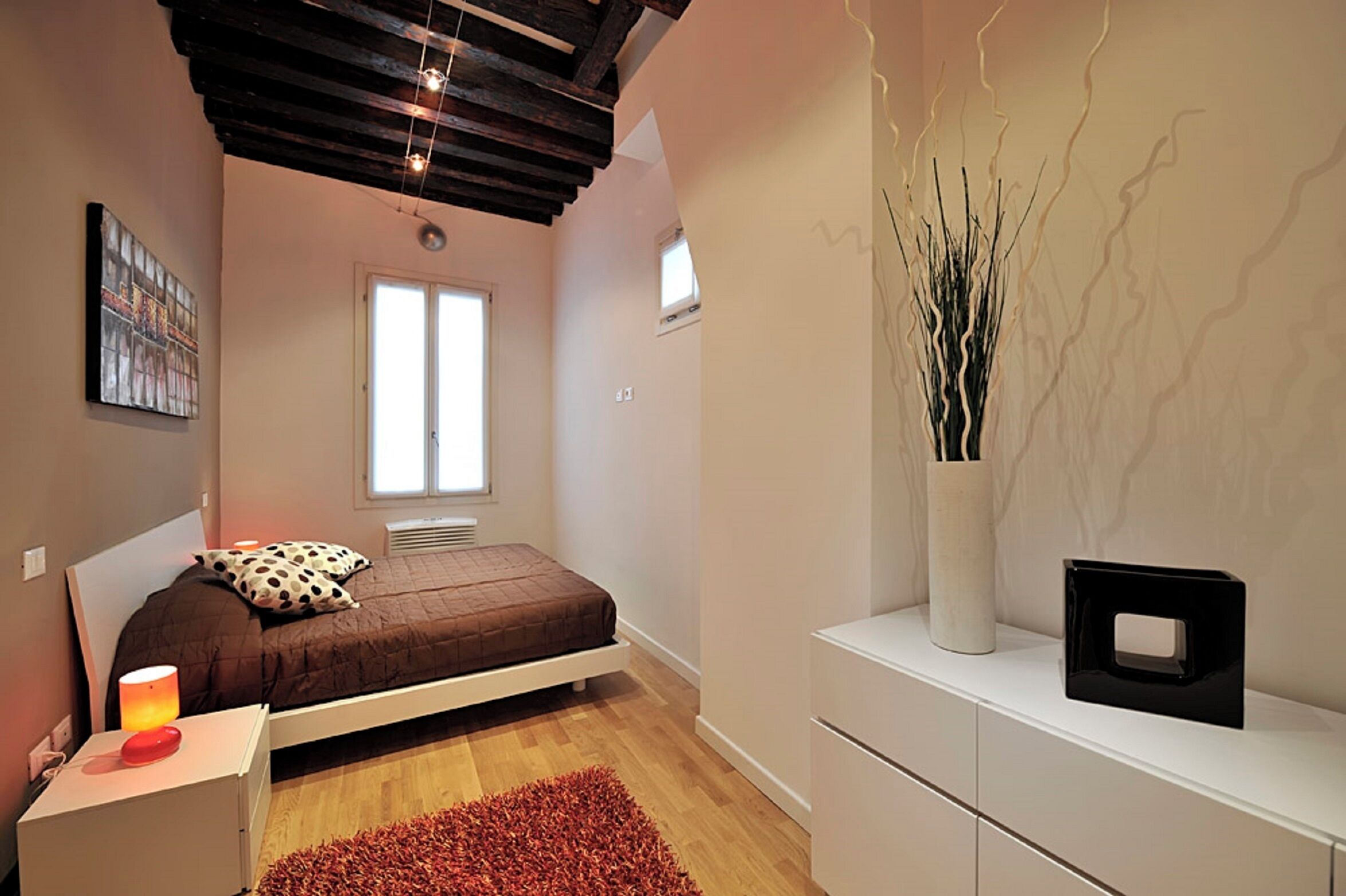 A bedroom with a bed, a nightstand, a red rug, a white dresser, and a vase with branches.