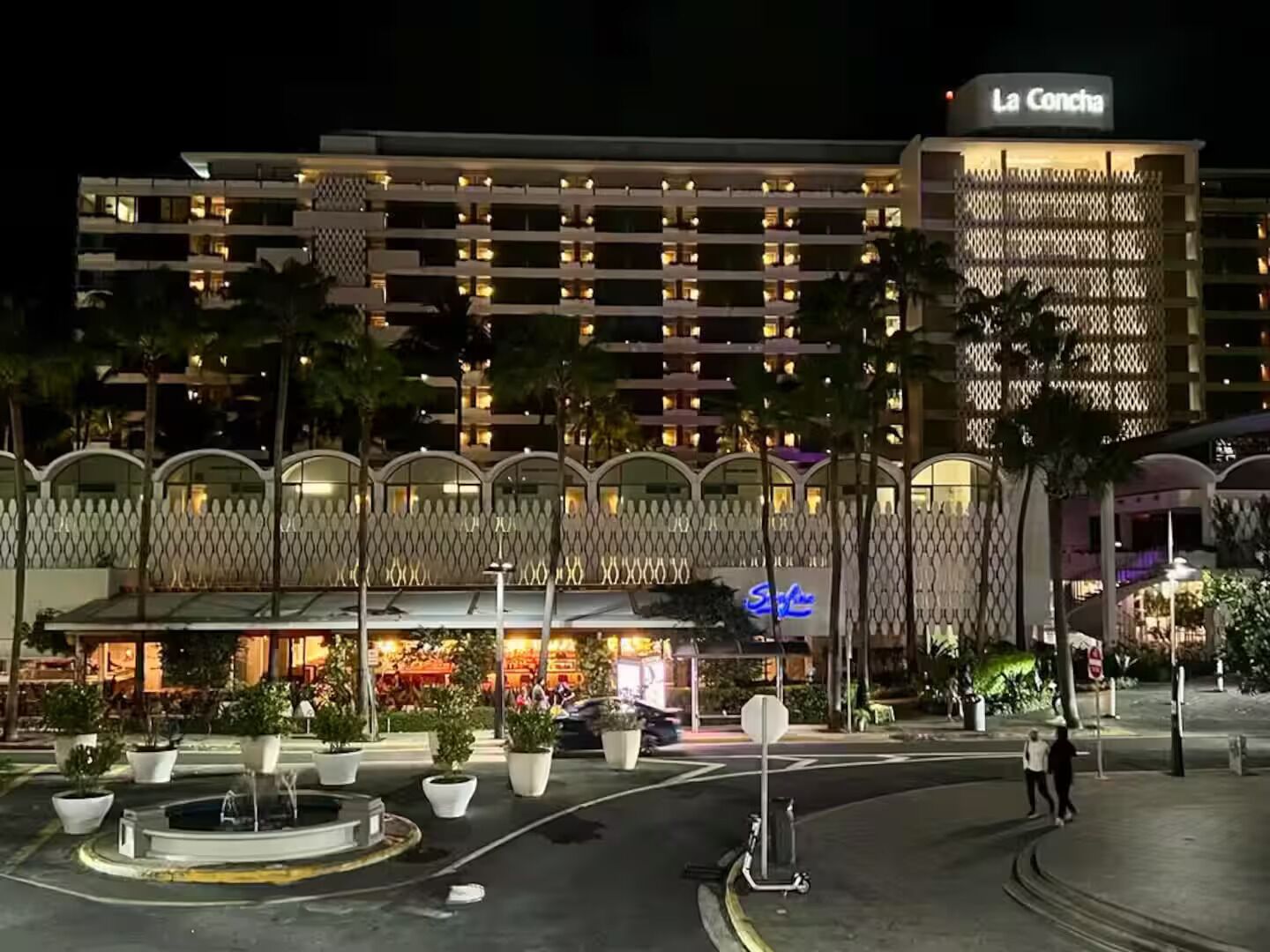 A multi-story hotel with a modern facade, a prominent sign reading "La Concha," and a landscaped courtyard with potted plants.
