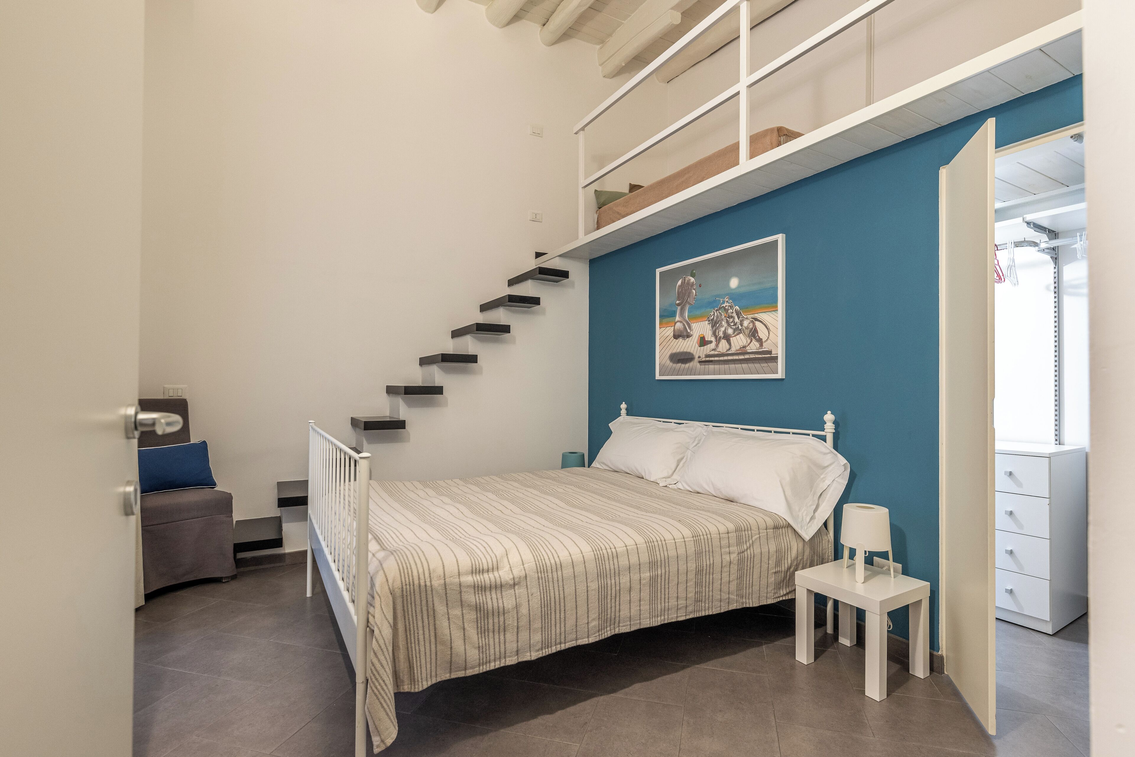 A bedroom with a bed, a nightstand, a chair, a painting on the wall, and a staircase.