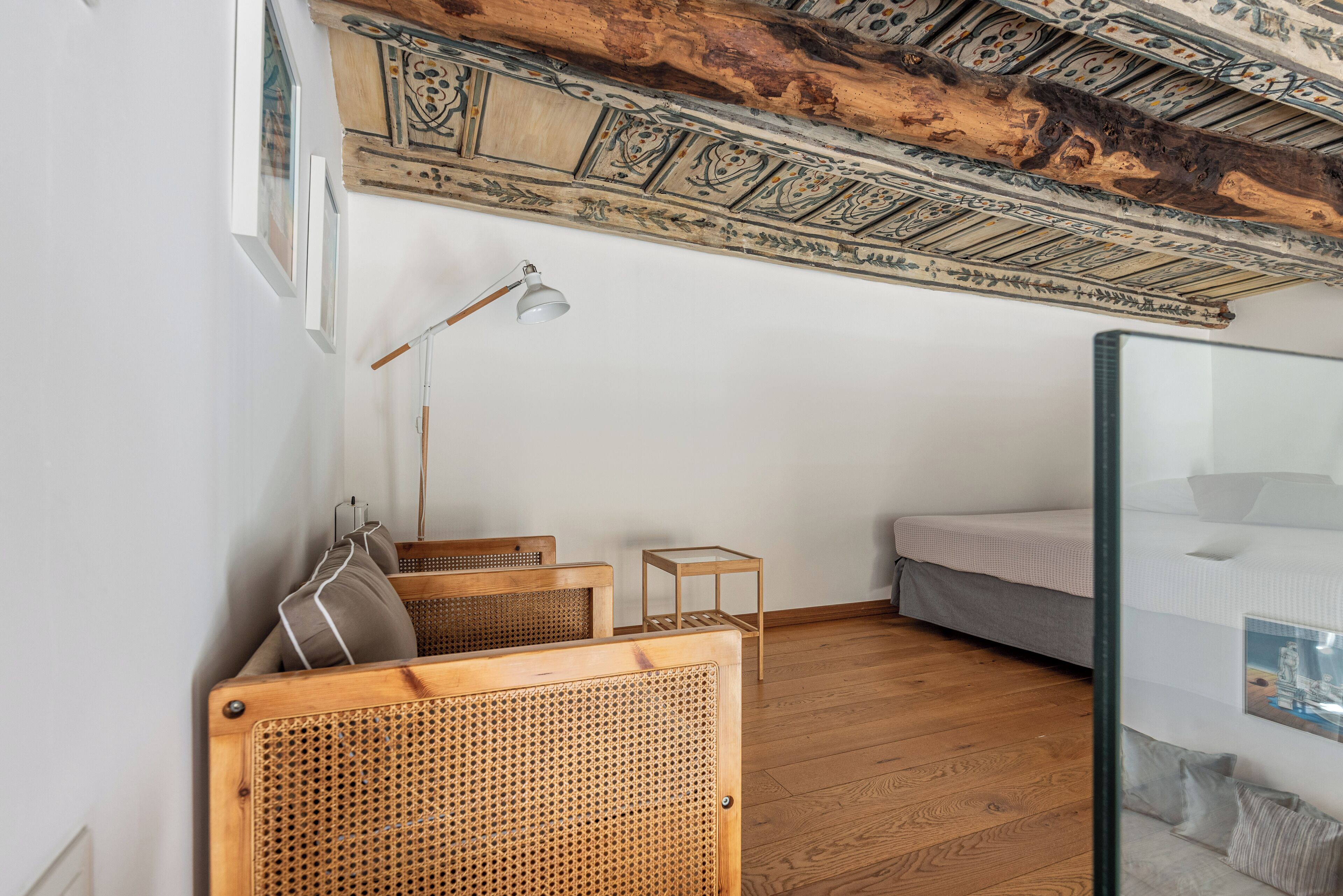 A room with a wooden ceiling, a bed, a rattan chair, a small table, and a floor lamp.