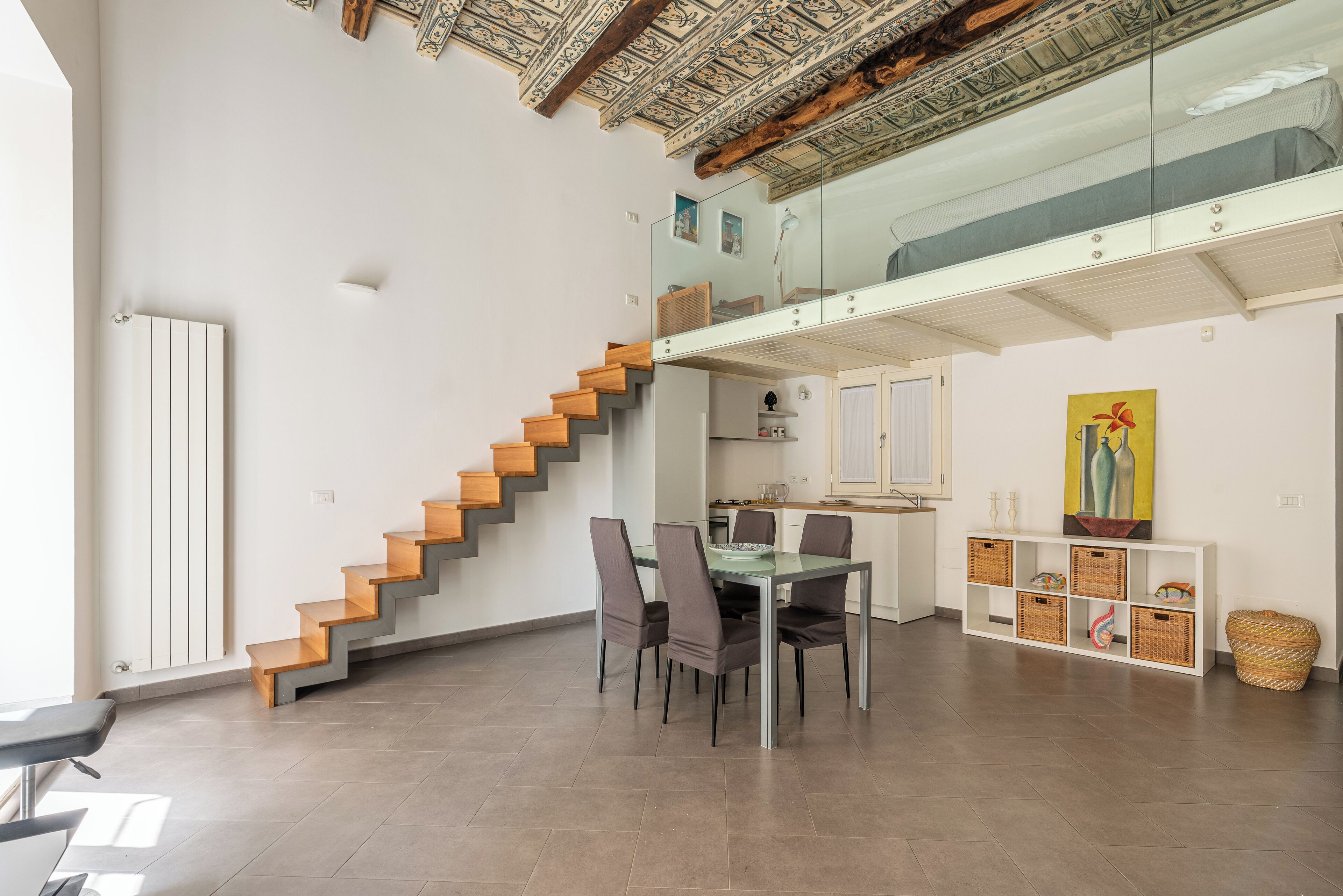 Um interior moderno com uma escada que leva a um mezanino, uma mesa de jantar com cadeiras e uma cozinha ao fundo.