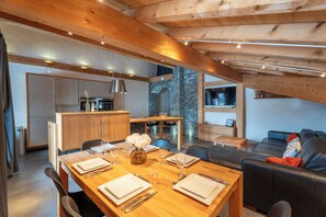 A modern kitchen with wooden beams, a dining area with a wooden table set for four, and a living area with a black leather sofa.