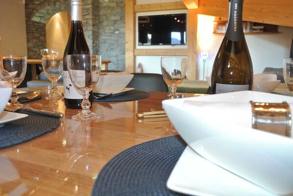 Una mesa de comedor con copas de vino, botellas y cubiertos, en una habitación con pared de piedra y un televisor al fondo.