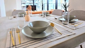 Ein Esstisch ist mit Tellern, Schüsseln, Gläsern und Besteck in einem modernen Raum mit großen Fenstern und Blick auf die Stadt gedeckt.