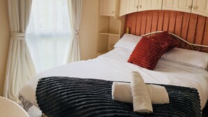 A neatly made bed with white and black bedding, a red pillow, and two folded towels. There are beige curtains, a wooden headboard, and a small shelf above the bed.