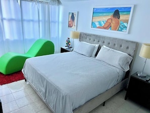 Un dormitorio ordenado con una cama, una silla verde y una obra de arte en la pared.