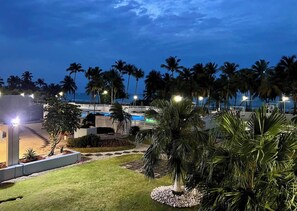 Zona de piscina en un resort, con palmeras, un puente y vista al océano al atardecer.