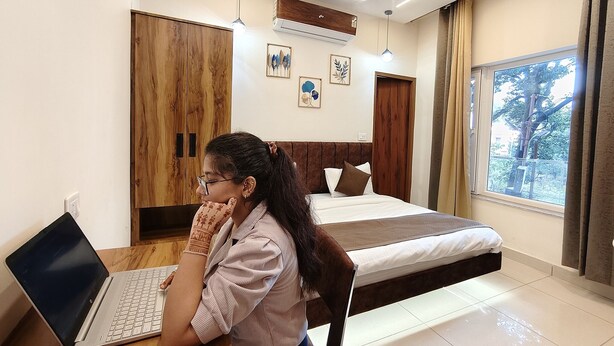 A bedroom with a bed, a desk with a laptop, and a window with a view of trees. - Hotel Ramayan inn (Rishikesh)
