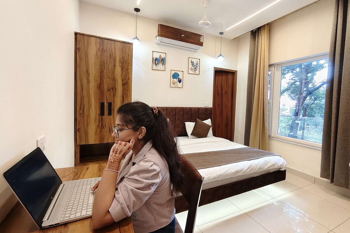 Un dormitorio con cama, un escritorio con una computadora portátil y una ventana con vista a árboles.