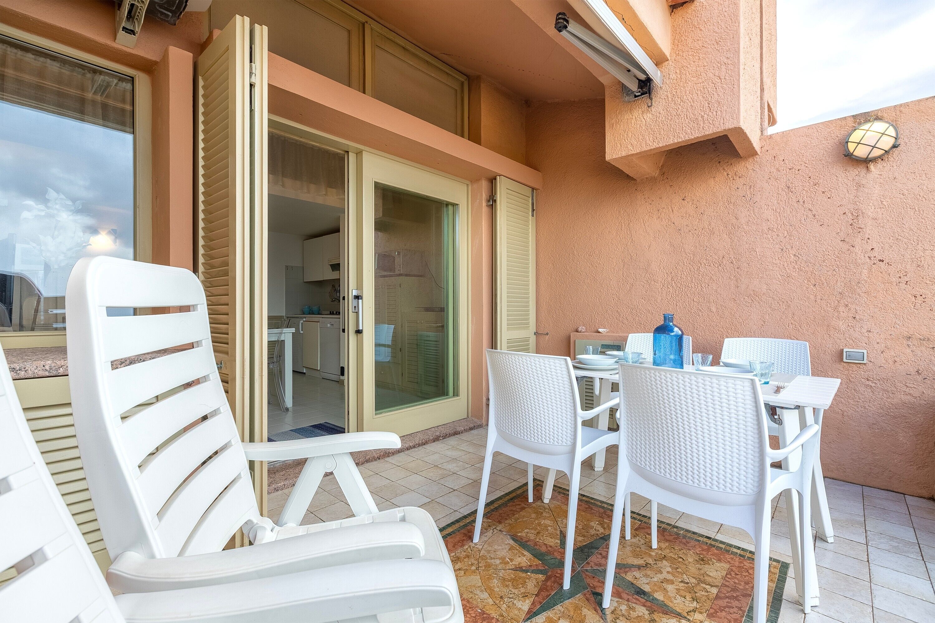 Un balcón con sillas de plástico blanco y una mesa, una puerta corrediza de vidrio y vista al interior.