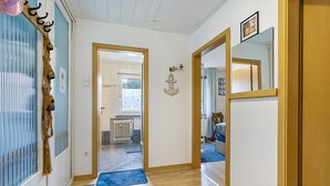 Un pasillo con suelo de madera, un felpudo azul, un adorno de ancla en la pared y una puerta que da a una habitación con alfombra azul y una silla.