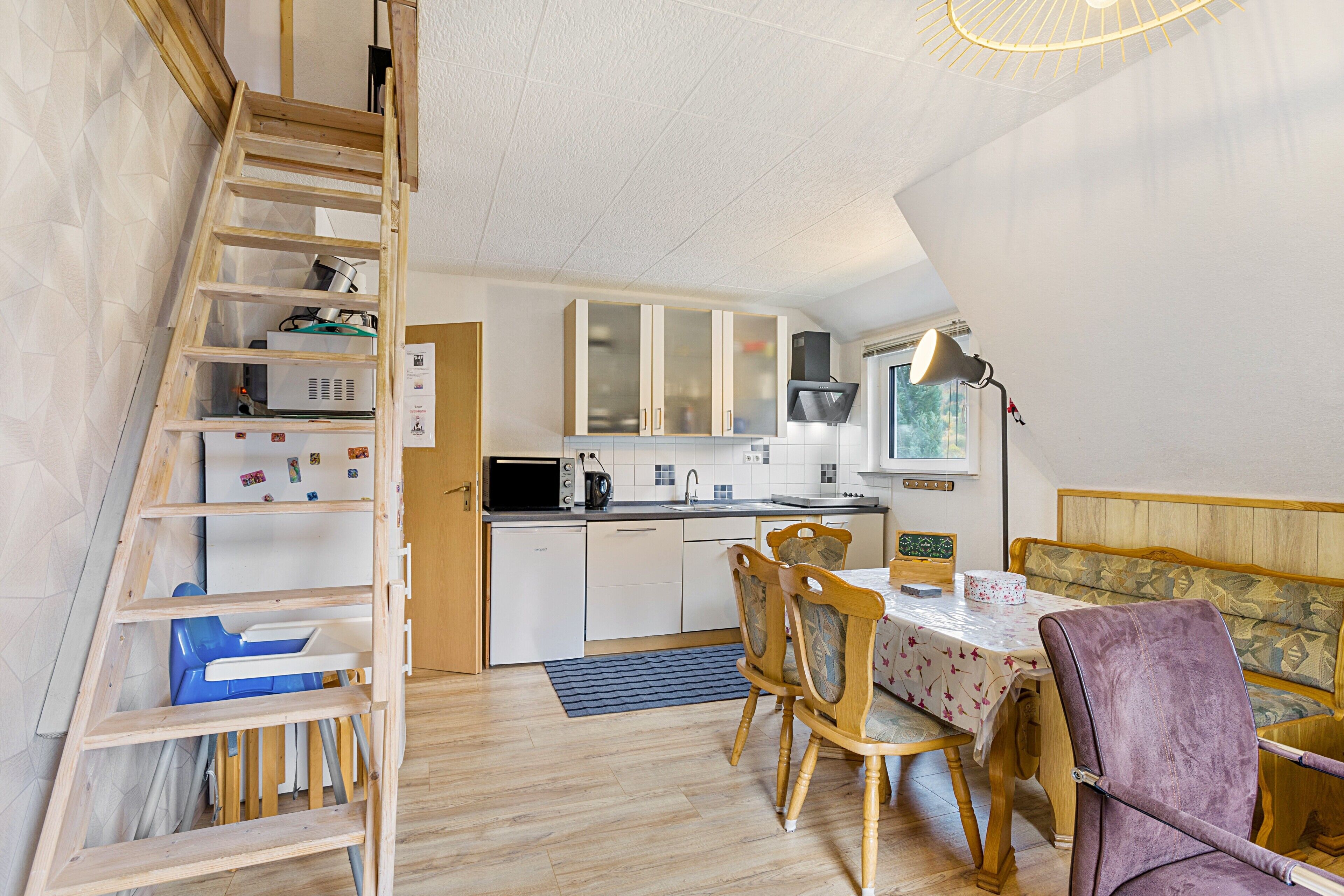 A compact kitchen with wooden cabinets, a dining table with chairs, a ladder, and a small seating area with a bench.