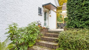 Un edificio blanco con una escalera de madera que conduce a una puerta, rodeado de vegetación y una pérgola.