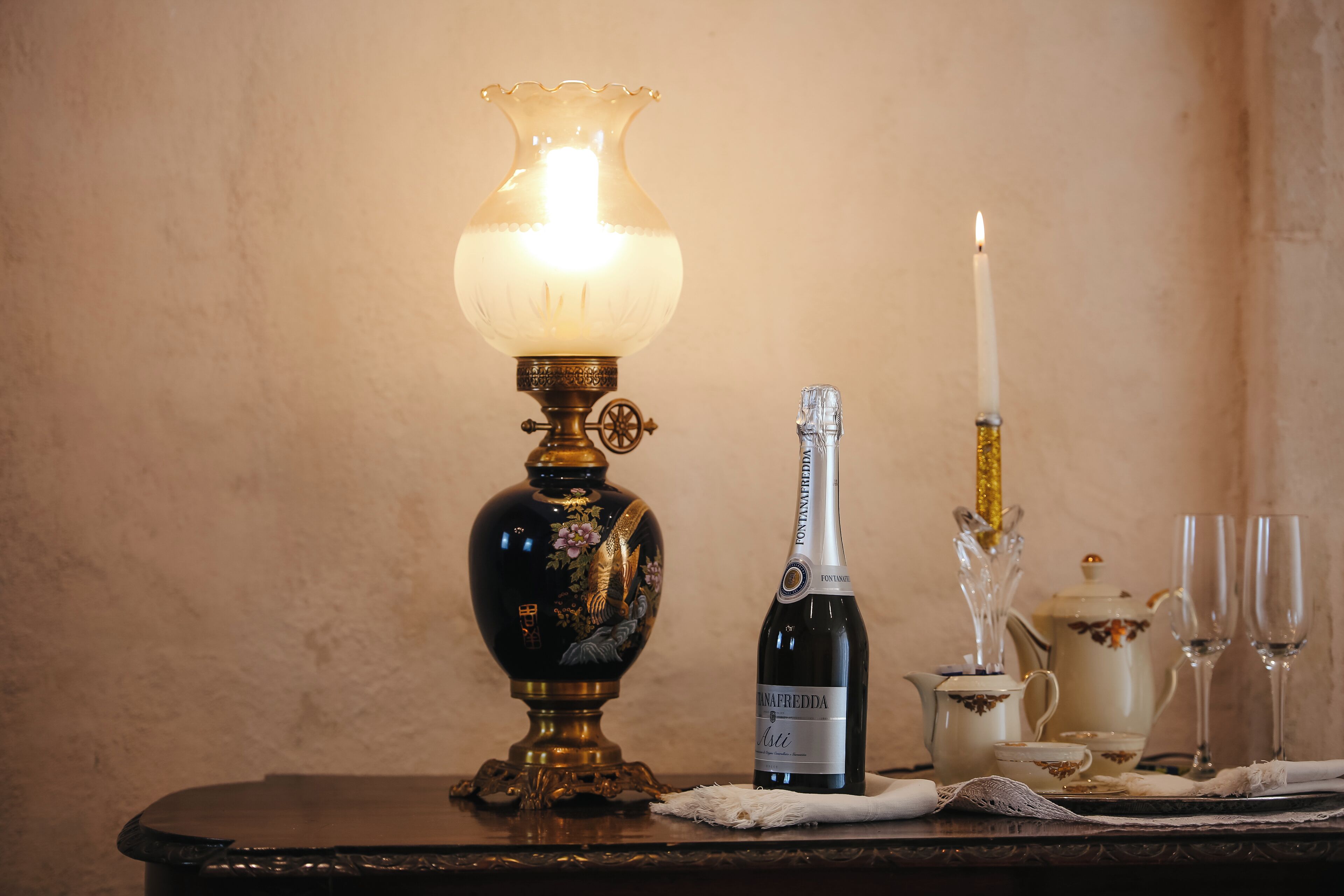 A table with a decorative lamp, a bottle of wine, a candle, and a teapot.
