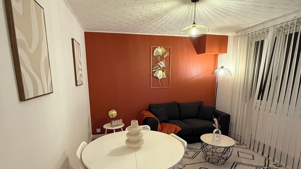 A modern living room with a white ceiling, orange walls, a black sofa, a white dining table with chairs, and a patterned rug. - Pleasant single-storey house in a quiet neighbourhood (Vierzon)