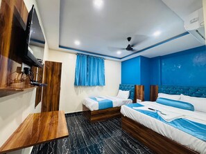 A hotel room with two beds, a TV mounted on the wall, a ceiling fan, and a wooden desk. - Welcome Airoli (Navi Mumbai)