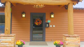 A wooden building with a gabled roof, a front door with a wreath, and two potted plants on either side.