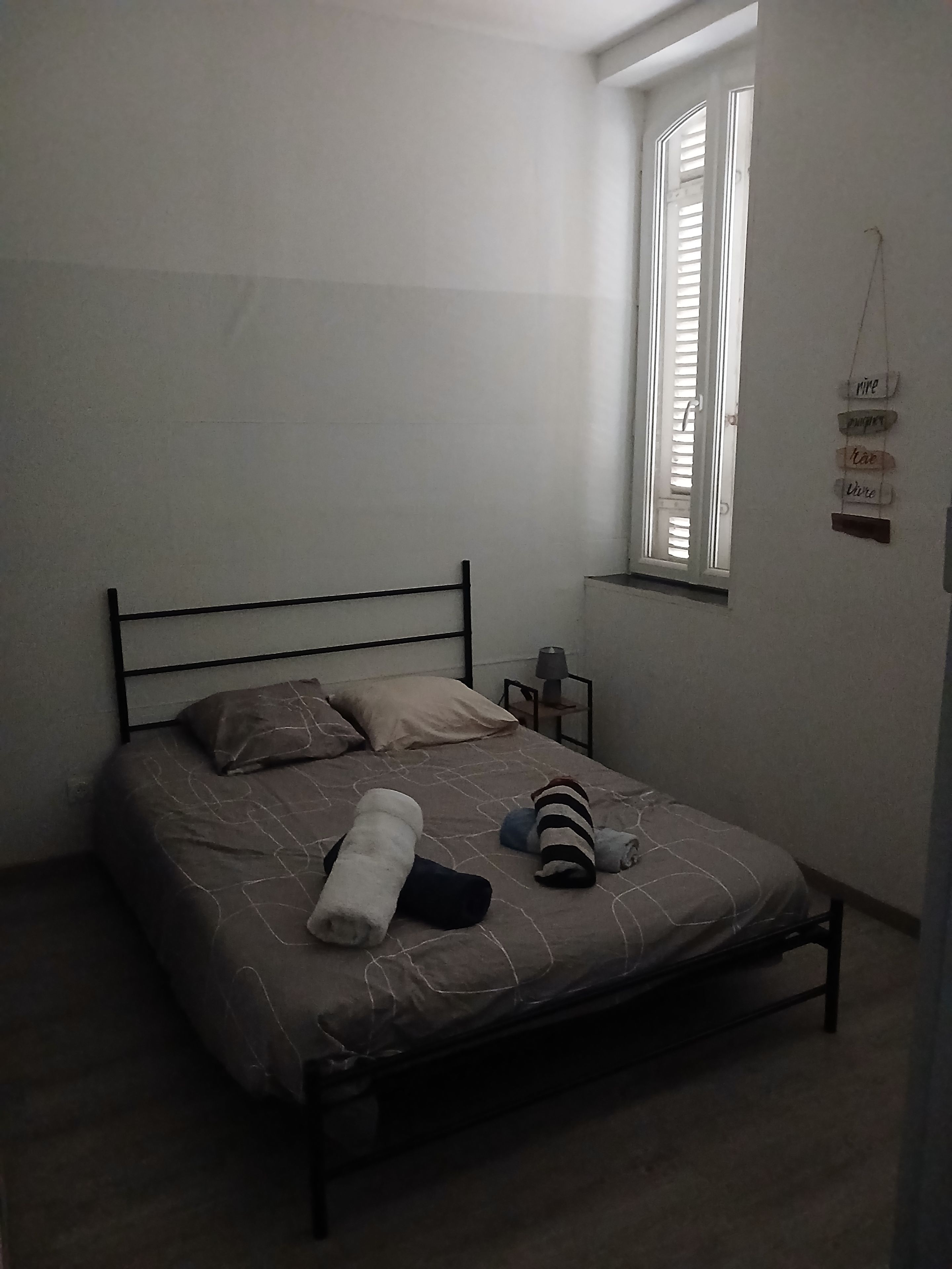 Een enkel bed met metalen frame, een raam met zonwering en een klein nachtkastje met een lamp.