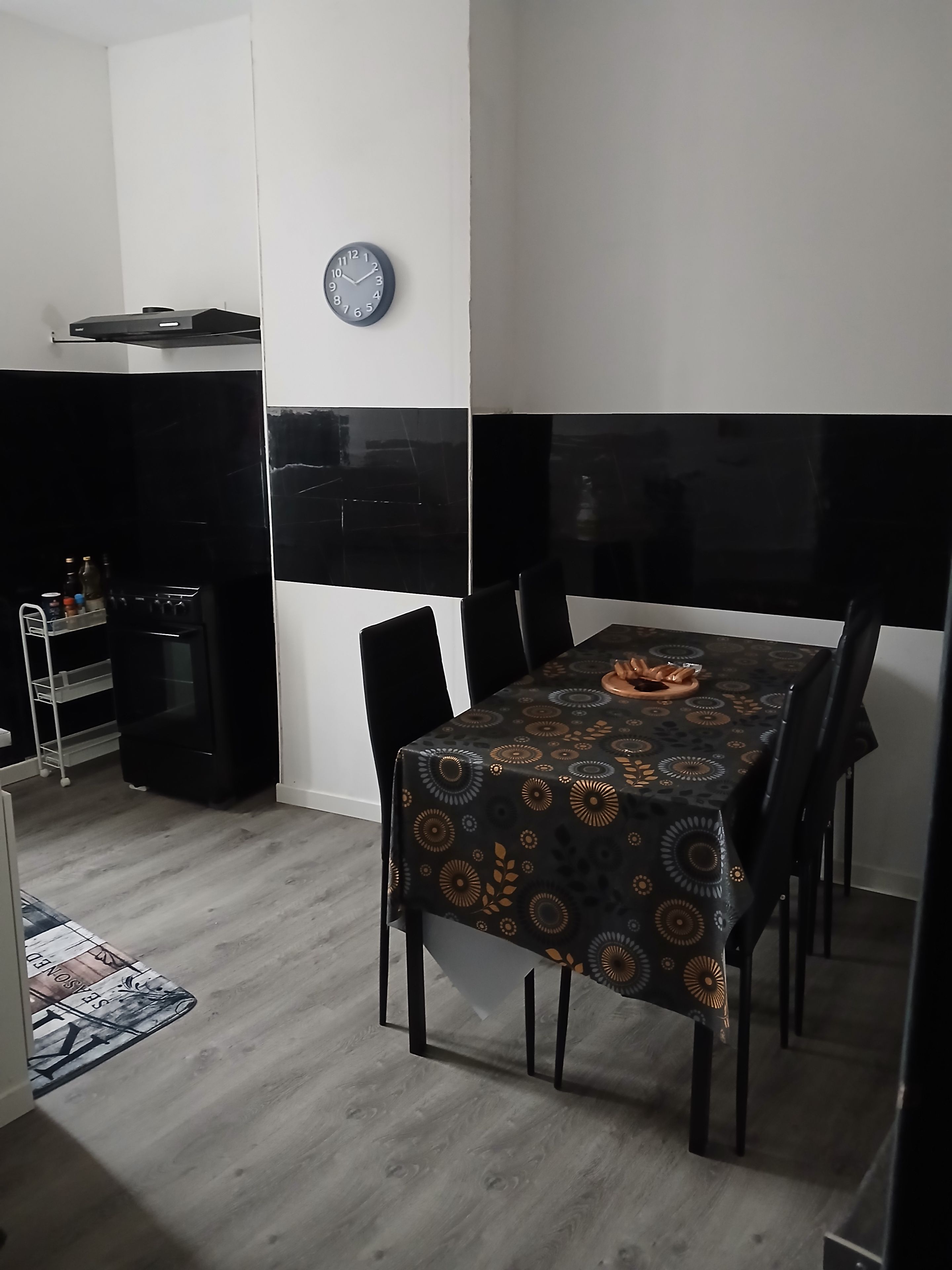 Een eetruimte met een tafel gedekt voor vier personen, een klok aan de muur, en een zwart-wit achterwand.