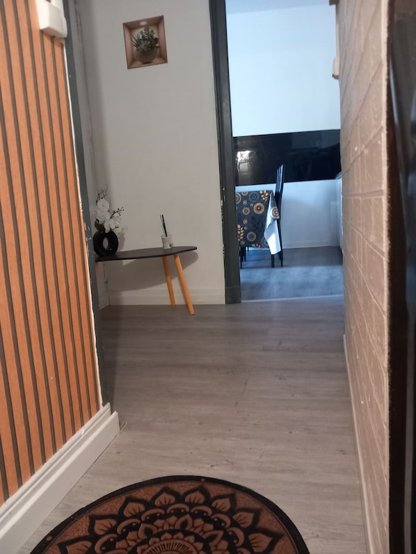 A hallway with a wooden floor, a small table with a vase of flowers, and a view into a room with a patterned tablecloth.
