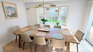 Un coin repas avec une table en bois, six chaises, un lustre, un tableau et une vue sur l’extérieur.