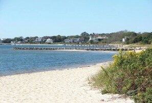 Ein Strand mit sandigem Ufer, einer in das Wasser ragenden Mole und Häusern im Hintergrund.