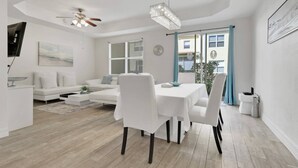 Ein modernes Wohnzimmer mit Essbereich, weißen Möbeln und einem großen Fenster mit blauen Vorhängen.