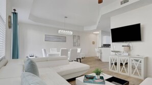 Ein modernes Wohnzimmer mit einem weißen Sofa, einem Couchtisch mit Dekoration, einem Essbereich mit weißen Stühlen und einer Küche im Hintergrund.